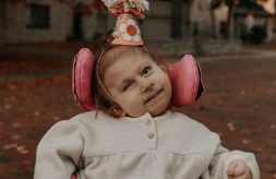 Eloise is wearing a light grey, long-sleeve fleece top and a cone-shipped birthday hat. The hat is made of light orange paper and that is decorated with a peace sign, flowers, a smiley face and a pom-pom on top. Her wheelchair has bright pink support pads that really pop in this picture.