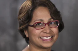 Dr. Shubhra Mukherjee has a beautiful smile, brown eyes and short, dark brown hair that frames her face. She is wearing glasses. The frames are dark red and are shaped like an oval that has been slightly squared on one end. She is wearing a navy colored, sleeveless top that has a honeycomb pattern woven into the fabric