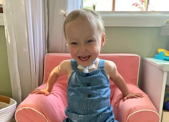 A young girl with a tracheostomy tube smiles at the camera while sitting in a small pink chair
