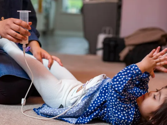 A mother feeds her daughter through a feeding tube in her abdomen as the young girl lies on her back