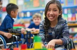 A girl with dark skin sits in a wheelchair and smiles as she plays with blocks inside an inclusive classroom for children with physical and intellectual disabilities.