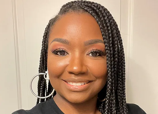 Meeka Harris earns the 2024 DSCC Award of Merit. She has long, dark hair that is parted at the side and braided in many narrow braids, She has light brown skin, a beautiful smile and brown eyes, She is wearing a grey t-shirt and silver hoop earrings.