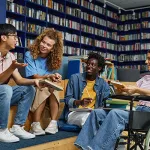 A diverse group of four students, including a young woman in a wheelchair, gathered in a college library and enjoying discussion