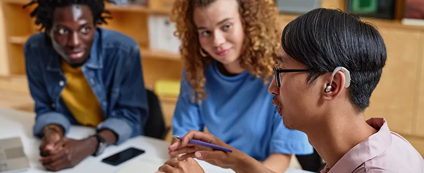 A young Asian man wearing a hearing aid device speaks with two other college-age students in a campus library space