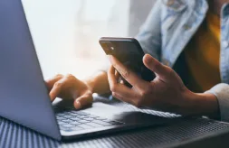 Close up of a set of dark-skinned hands with one hand holding a cell phone while the other hand types on a laptop