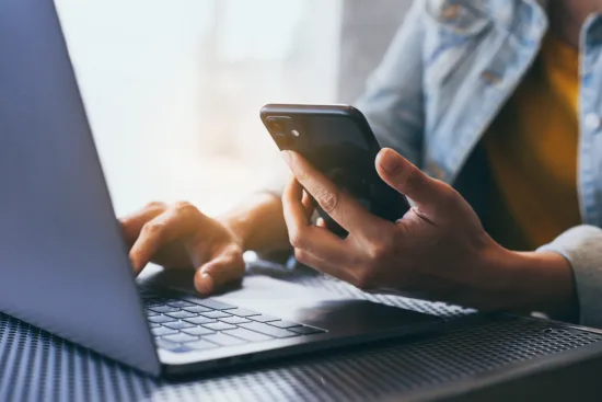 Close up of a set of dark-skinned hands with one hand holding a cell phone while the other hand types on a laptop