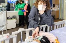 A woman in a mask checking the tracheostomy tube of an infant medical training mannequin in a simulation room setting