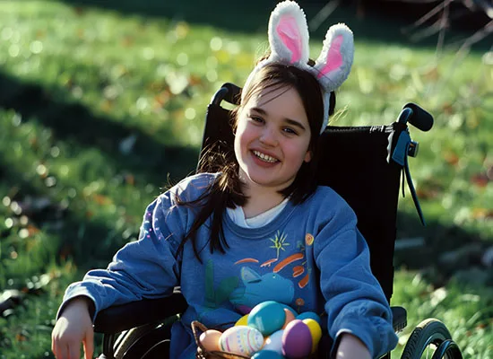 A girl wearing bunny ears has a basket full of colorful eggs on her lap. She is smiling in her wheelchair.