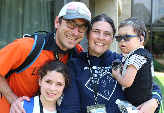 A family of four smiles together in front of the Illinois School for the Deaf. The mom and dad are in the center of the photo. The mom holds her four-year-old son. He wears glasses and has hearing aids. The dad also has glasses. He puts his hand on the shoulder of his seven-year-old daughter.